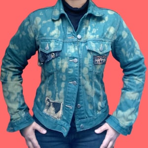 Puede incluir: Una chaqueta vaquera verde azulado con un patrón decolorado y botones plateados. La chaqueta tiene dos bolsillos en el pecho y parches con texto e imágenes. Las mangas son largas y el cuello es de estilo clásico. La chaqueta se lleva sobre una camiseta negra.