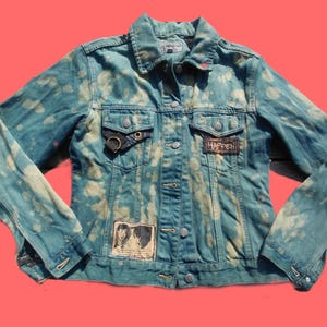 May include: A distressed denim jacket in shades of blue with bleach-like spots. The jacket features a button-up front, pockets, and patches with the word "HAPPEN" and a black and white image. A black ring detail is on the left pocket.