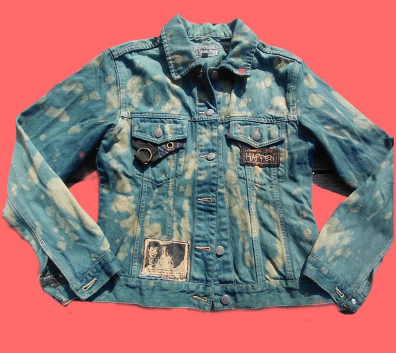 May include: A distressed denim jacket in shades of blue with bleach-like spots. The jacket features a button-up front, pockets, and patches with the word "HAPPEN" and a black and white image. A black ring detail is on the left pocket.