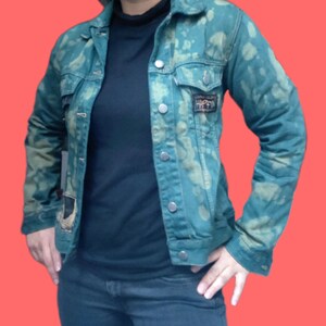 May include: Teal denim jacket with a bleached pattern and silver buttons. The jacket has two chest pockets and a small patch on the right side. The model is wearing a black turtleneck and dark blue jeans.