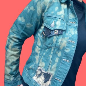 May include: Teal denim jacket with a unique bleached pattern. Features a button-up front, chest pockets, and a patch with a graphic design. The jacket has a metal ring detail and a small plaid accent on the pocket.