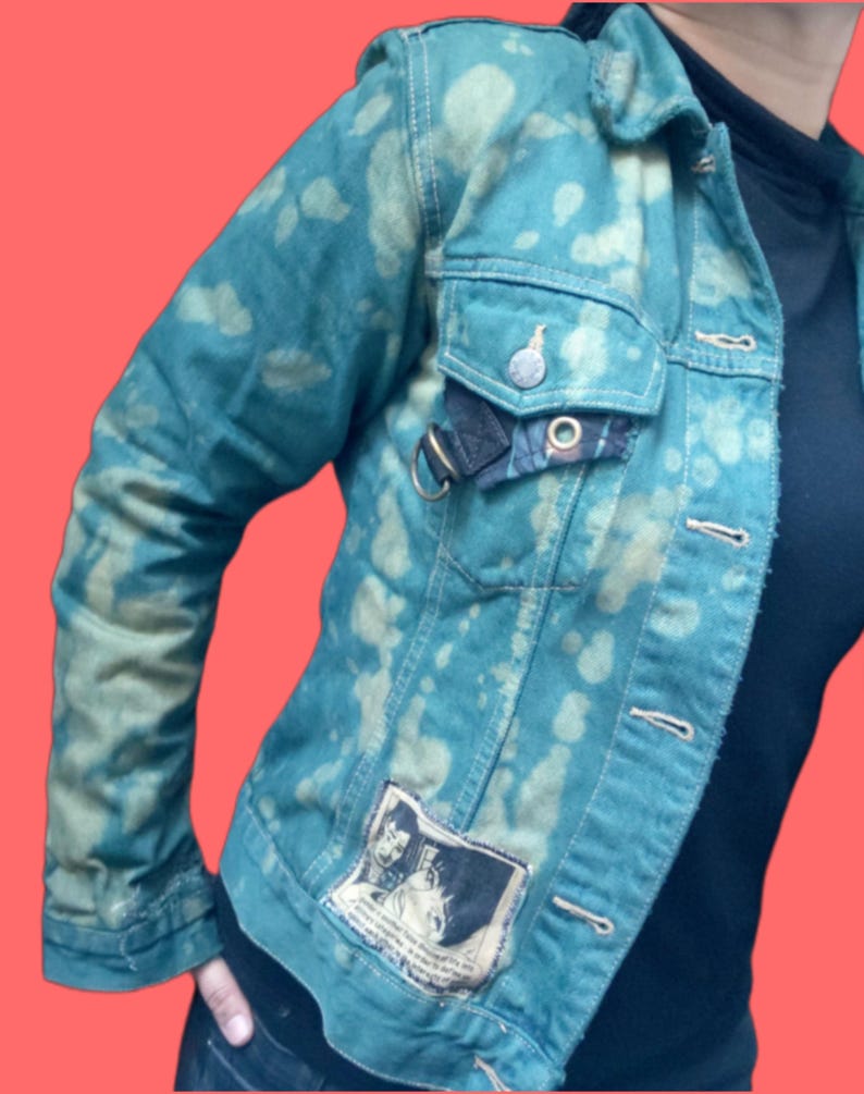 May include: Teal denim jacket with a unique bleached pattern. Features a button-up front, chest pockets, and a patch with a graphic design. The jacket has a metal ring detail and a small plaid accent on the pocket.