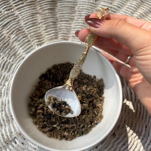 Pode incluir: Uma tigela de cerâmica branca cheia de ervas secas, com uma colher decorativa dentro. A colher tem um cabo dourado com um design enrolado em arame e uma pequena pedra de cor clara. A tigela da colher é branca e em forma de concha.