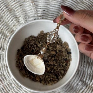 May include: A handcrafted spoon with a seashell bowl and a decorative gold-colored handle. The handle is adorned with clear crystals and a pearl-like bead. The spoon is held over a white bowl filled with dried herbs.