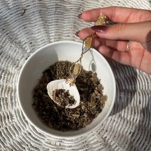 Pode incluir: Um pingente de arame dourado com pedras amarelas e transparentes é segurado acima de uma tigela branca cheia de ervas secas. O pingente está conectado a um anel com uma pedra correspondente. A tigela está sobre uma superfície branca tecida.