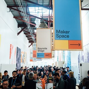 May include: An interior shot of a crowded event space with signs hanging from the ceiling. Signs indicate "Maker Space" and "Keynote Stage". People are gathered in the foreground, with a focus on the event's atmosphere and signage.