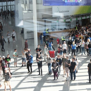 May include: A large, bright, modern convention center interior filled with people. Many individuals are walking around, some carrying bags or wearing backpacks. Signage for "#GAMING" and "koelnmesse" is visible.