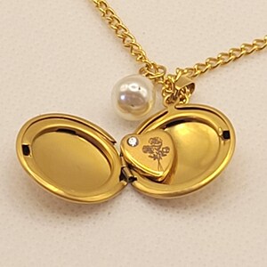 Puede incluir: Collar con colgante dorado, cadena, perla y un dije en forma de corazón en el interior. El colgante está abierto, revelando un pequeño dije de corazón con un diseño floral y un pequeño cristal. El collar está sobre una superficie blanca.