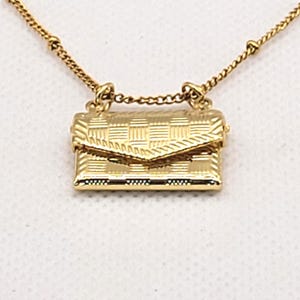 Op de afbeelding: Een goudkleurige ketting met een kleine tasvormige hanger. De hanger heeft een getextureerd, geweven patroon en een sluiting. De ketting heeft een delicate goudkleur, met een kleine ketting en een kralenontwerp.