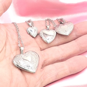 May include: Four silver heart-shaped locket necklaces with detailed floral patterns. Each locket is suspended from a fine silver chain. The lockets are shown on a pink satin surface.