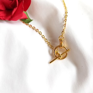 Könnte beinhalten: Eine goldene Kettenkette mit einem Toggle-Verschluss. Der Verschluss ist ein runder Ring mit einer Stange, die durch ihn gleitet.