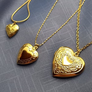 Könnte beinhalten: Drei goldfarbene herzförmige Medaillons an zarten Goldketten. Jedes Medaillon weist ein ausgeprägtes florales oder geschwungenes Prägemuster auf seiner Oberfläche auf. Die Medaillons variieren in der Größe, mit einem kleinen, mittleren und großen Herzanhänger.