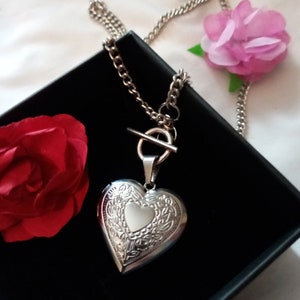 Può includere: Una collana con un ciondolo a forma di cuore in argento con un design floreale dettagliato. La collana ha una chiusura a levetta ed è esposta su uno sfondo nero.