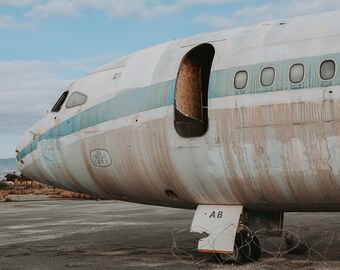 May include: A weathered, white airplane fuselage with a blue stripe and several windows. The airplane is sitting on a tarmac with a barbed wire fence in the foreground. The tail of the plane has the letters "AB" painted on it.