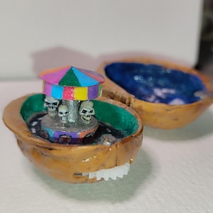 Op de afbeelding: Een miniatuurcarrousel met een kleurrijk gestreept baldakijn en vier schedelfiguren, gemonteerd in een gehalveerde walnootschelp. De schelp is geschilderd met een groen en blauw interieur.