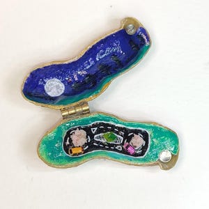Puede incluir: Una pequeña caja de baratijas en forma de concha abierta. La mitad superior representa una escena nocturna con cielo azul, árboles oscuros y luna llena. La mitad inferior muestra una carretera con un coche amarillo y rosa. La caja está adornada en dorado.