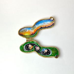 Puede incluir: Caja de baratijas pintada a mano, abierta para revelar un paisaje y una carretera con coches en miniatura. La caja tiene un borde dorado y está articulada. La sección superior muestra una puesta de sol. Sobre fondo blanco.