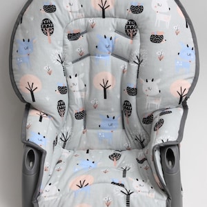 May include: A gray baby seat insert with a whimsical pattern of cartoon deer, trees, and hedgehogs in pastel colors. The seat has a padded design and a dark gray trim, with openings for straps and a secure fit.