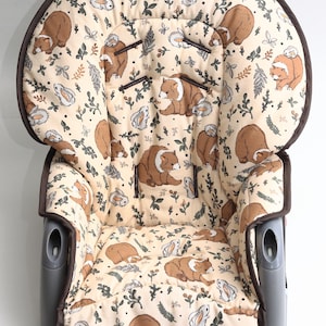 Puede incluir: Inserto para trona beige con ribete marrón y un estampado repetido de osos marrones, conejos blancos y follaje verde. El inserto tiene un diseño acolchado y está hecho de tela suave.