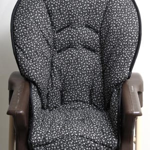 May include: A high chair seat cushion with a black background and a white floral pattern. The cushion is padded and has a black trim. The high chair frame is brown and beige.