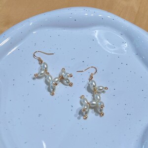 May include: A pair of gold earrings with white pearl beads shaped like a balloon animal. The earrings are on a white speckled surface.