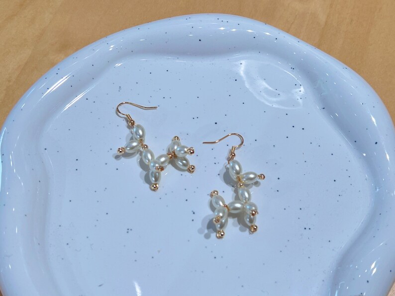 May include: A pair of gold earrings with white pearl beads shaped like a balloon animal. The earrings are on a white speckled surface.