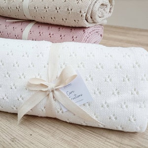May include: A white, crocheted baby blanket with a diamond pattern. The blanket is tied with a natural-colored ribbon and a tag that reads "Care Instructions".