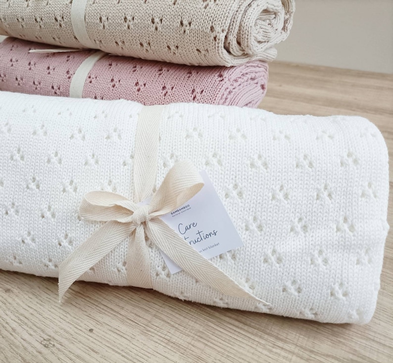 May include: A white, crocheted baby blanket with a diamond pattern. The blanket is tied with a natural-colored ribbon and a tag that reads "Care Instructions".