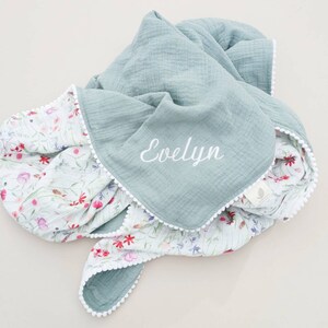 May include: A soft, light blue and white muslin baby blanket with a floral print on one side and the name "Evelyn" embroidered on the other side. The blanket has a white pom-pom trim.