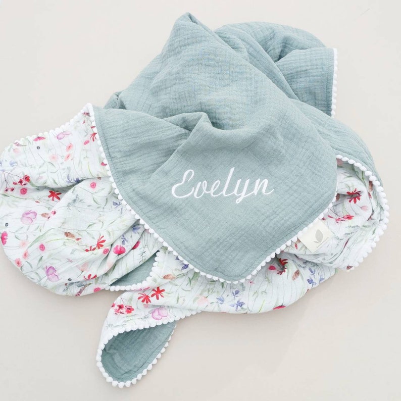 May include: A soft, light blue and white muslin baby blanket with a floral print on one side and the name "Evelyn" embroidered on the other side. The blanket has a white pom-pom trim.