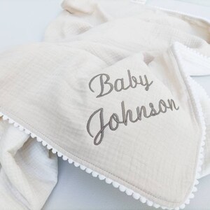 May include: A cream-colored baby blanket with a white pom-pom trim and the words "Baby Johnson" embroidered in gray.