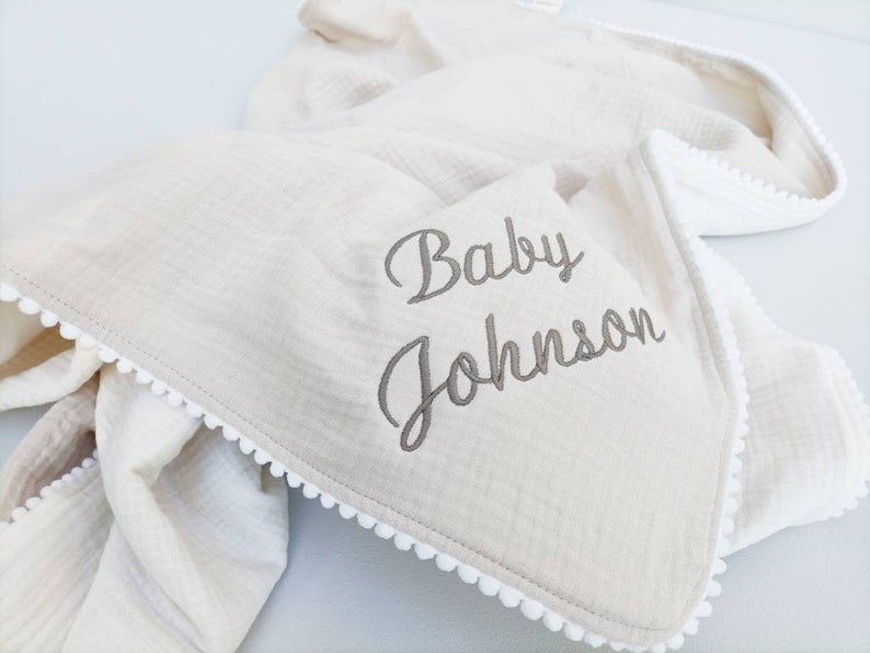 May include: A cream-colored baby blanket with a white pom-pom trim and the words "Baby Johnson" embroidered in gray.