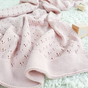 May include: A soft pink knitted baby blanket with a delicate openwork pattern. The blanket is folded and draped over a white fluffy surface.