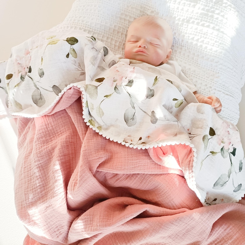 May include: A baby blanket with a floral pattern and a solid pink backing. The top layer features a white background with pink and green floral designs. The blanket has a scalloped edge with white pom-poms. The bottom layer is a solid, soft pink.