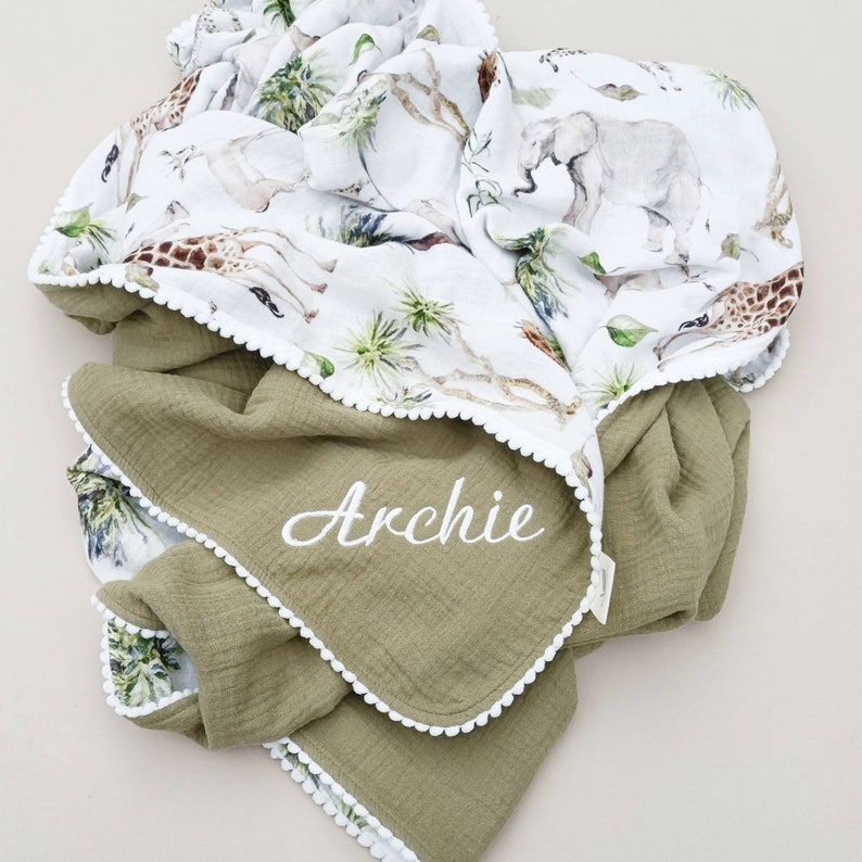 May include: A green muslin baby blanket with white pom pom trim and a white embroidered name "Archie". The blanket is reversible and the other side features a white cotton fabric with a jungle animal print.