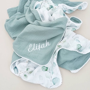 May include: A soft, light blue muslin baby blanket with a white pom-pom trim and a white floral print on one side. The blanket is personalized with the name "Elijah" embroidered in white on the solid side.