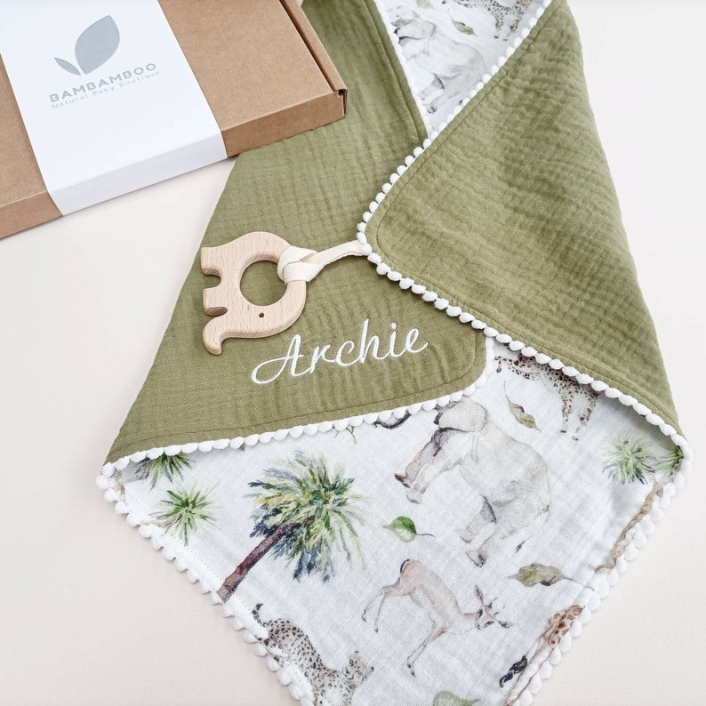 Op de afbeelding: Een babydeken met een groene bovenlaag en een witte onderkant met een patroon van olifanten en andere dieren. De deken heeft witte pompom-randen en de naam "Archie" is in het wit geborduurd. Er is een houten bijtring bevestigd.