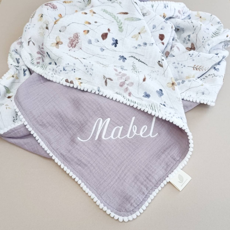 May include: A reversible baby blanket with a floral pattern on one side and a solid lavender color on the other. The blanket is trimmed with white pom-poms and features the name "Mabel" embroidered in white.
