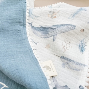 Op de afbeelding: Een babydeken met een blauw-witte walvisprint. De deken heeft aan de ene kant een blauwe, getextureerde stof en aan de andere kant een witte stof met walvissen, narwallen en zeegroei. De randen zijn afgewerkt met witte pompons.