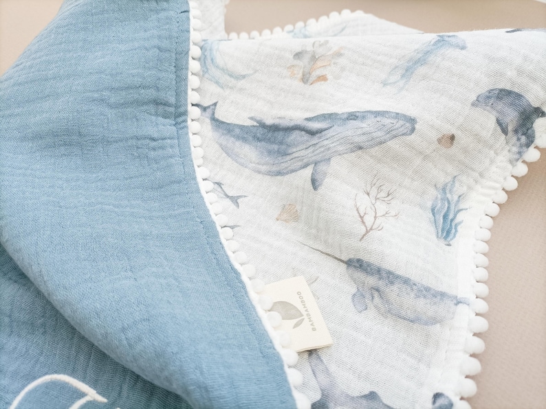 Op de afbeelding: Een babydeken met een blauw-witte walvisprint. De deken heeft aan de ene kant een blauwe, getextureerde stof en aan de andere kant een witte stof met walvissen, narwallen en zeegroei. De randen zijn afgewerkt met witte pompons.