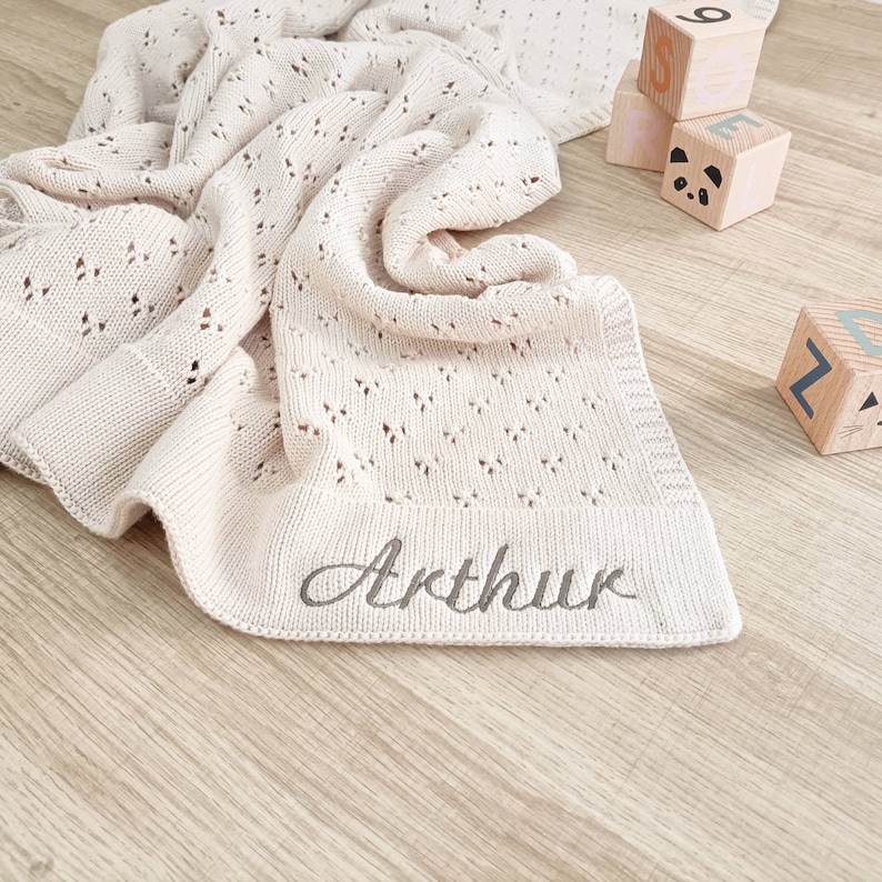 May include: A cream-colored knitted baby blanket with a delicate openwork pattern. The blanket is personalized with the name "Arthur" embroidered in a cursive font.