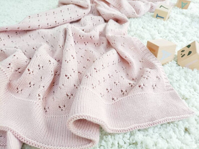 May include: A soft, light pink knitted baby blanket with a delicate openwork pattern. The blanket is draped over a white, fluffy surface. Wooden blocks with letters and shapes are in the background.