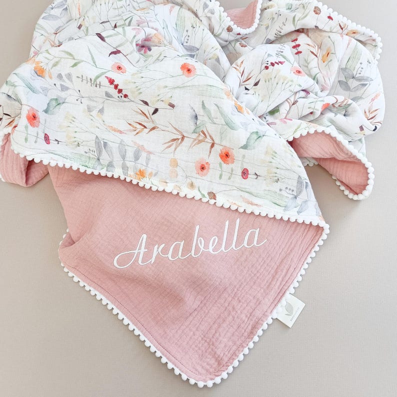 May include: A reversible baby blanket with a floral pattern on one side and a solid pink color on the other. The blanket is trimmed with white pom-poms and features the name "Arabella" embroidered in white.