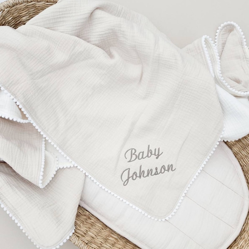 May include: A beige muslin baby blanket with white pom-pom trim and the embroidered text "Baby Johnson".