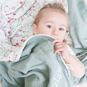 May include: A baby lies nestled in a soft, sage green blanket with a white pom-pom trim. The blanket is personalized with the name "Evelyn" in white script. A floral patterned blanket with pink and red flowers is visible in the background.