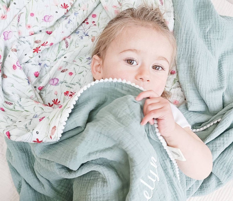 May include: A baby lies nestled in a soft, sage green blanket with a white pom-pom trim. The blanket is personalized with the name "Evelyn" in white script. A floral patterned blanket with pink and red flowers is visible in the background.