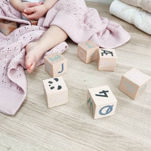 May include: Wooden alphabet blocks with pastel letters and a panda face are scattered on a light wood surface. A soft, pink, open-weave blanket and a child's legs are in the frame. The blocks are a classic toy.