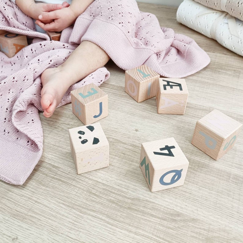 May include: Wooden alphabet blocks with pastel letters and a panda face are scattered on a light wood surface. A soft, pink, open-weave blanket and a child's legs are in the frame. The blocks are a classic toy.