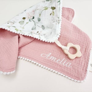 Op de afbeelding: Een roze babydeken met witte pompom-afwerking en een bloemenprint op de achterkant. De deken heeft de naam "Amelia" in het wit geborduurd. Een houten bloemvormige bijtring is bevestigd.