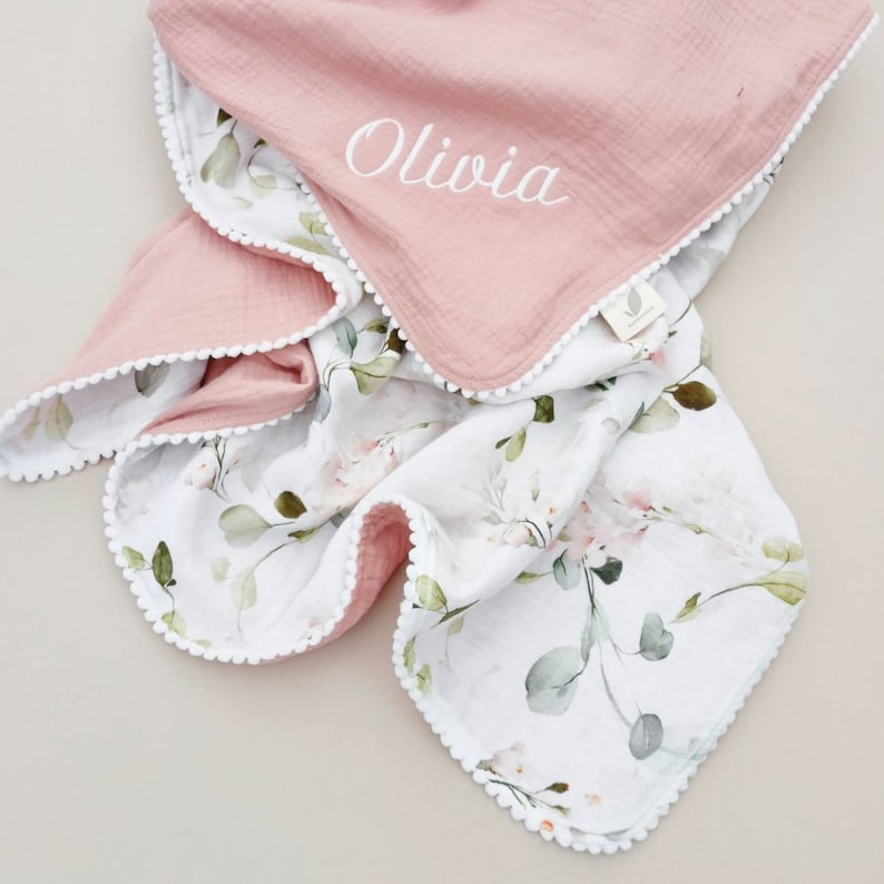 May include: A pink and white muslin baby blanket with a floral pattern and white pom-pom trim. The blanket is personalized with the name "Olivia".
